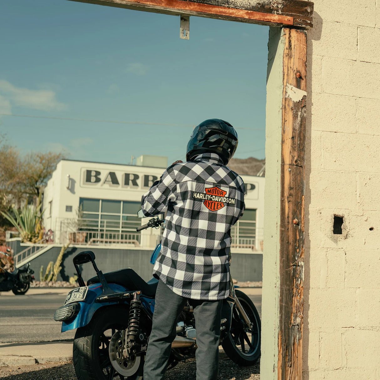 Person wearing a checkered shirt with a Harley-Davidson logo next to a blue motorcycle, with a building in the background.